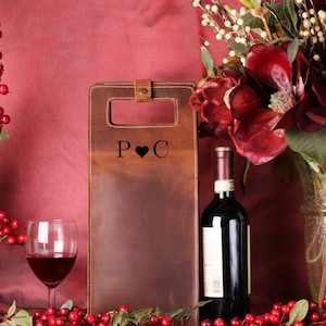 May include: A brown leather wine bottle carrier with the initials "P ❤️ C" is displayed next to a wine glass filled with red wine and a bottle of red wine. The background features red berries and flowers.