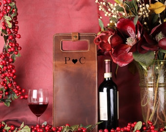 May include: A brown leather wine bottle carrier with the initials "P ❤️ C" is displayed next to a wine glass filled with red wine and a bottle of red wine. The background features red berries and flowers.