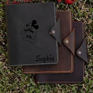 May include: Three leather-bound journals with decorative stitching. The top journal is black and features a Mickey Mouse illustration and the name "Sophia" engraved on the front. The middle journal is brown and the bottom journal is dark brown.