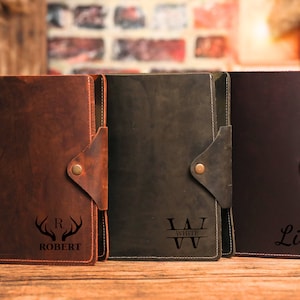 May include: Three leather journals in shades of brown, green, and burgundy. Each journal has a flap closure with a brass button. The first journal is embossed with antlers and the name "ROBERT". The second journal has the name "WHITE". The third journal has the name "Lily".