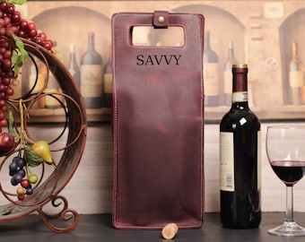 Può includere: Un portabottiglie da vino in pelle color bordeaux scuro di forma rettangolare, dotato di manico superiore con chiusura a pressione color ottone e la parola SAVVY impressa in lettere nere sulla parte anteriore, con cuciture bianche sui bordi.