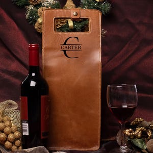 May include: A tan leather wine bottle carrier featuring a rectangular cutout handle with a snap closure and the black printed text CARTER centered within a stylized letter C. The carrier is positioned next to a wine bottle and a glass of red wine.