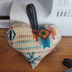 May include: A heart-shaped fabric ornament with a patterned design in beige, teal, red, and brown. It features a black ribbon loop and a wooden button. The ornament is likely handmade and could be used as a decorative item.