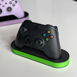 May include: Two video game controllers, one purple and one black, are displayed on charging stands. The purple stand has a light purple base and the black stand has a green base.