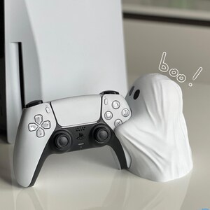 May include: A white PlayStation 5 controller with black buttons and a white ghost figurine with the word "boo!" written above it.
