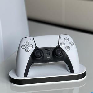 May include: A white PlayStation 5 DualSense controller on a white charging stand with a black trim. The controller has black buttons and a black PlayStation logo.
