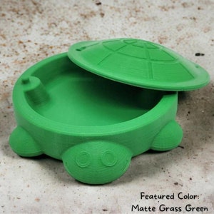 May include: A green turtle-shaped container with a lid. The container is made of plastic and has a smooth, matte finish. The lid has a textured design that resembles a turtle shell. The container is open and the lid is resting on top of it.