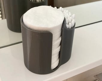 May include: A dark gray cylindrical bathroom organizer featuring a large front slot filled with a stack of white round cotton pads and a smaller side compartment containing a cluster of white cotton swabs for personal hygiene.