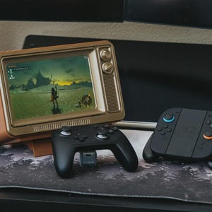 May include: A retro-style television displays a video game scene. A black controller and a black and blue game controller are in front of the TV. A black tablet is in the background.