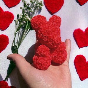 May include: A red 3D printed teddy bear shaped like a rose. The bear is being held in a person's hand. The background is a white fabric with red hearts.