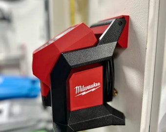May include: A red and black Milwaukee cordless inflator mounted on a white wall. The inflator has a black handle and base, with the Milwaukee logo in white. A black hose is coiled on the side.