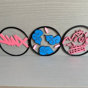 May include: Three black circular wall decor pieces with pink and blue designs. The first piece has pink diagonal lines, the second piece has blue clouds and pink pencils, and the third piece has a pink cartoon face.