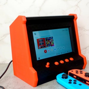 May include: A red and black arcade-style console with a screen displaying the Nintendo Switch menu. Two Nintendo Switch controllers are in the foreground, one blue and one red.