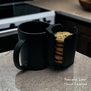 May include: A black ceramic mug with a built-in cookie holder. The holder is filled with chocolate chip cookies. The mug and cookie holder are both matte charcoal in color.  Featured Color: Matte Charcoal