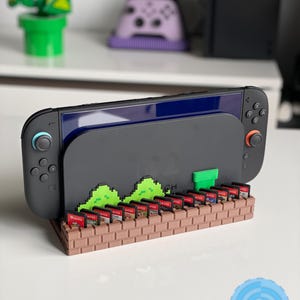 May include: A black Nintendo Switch console sits on a brown brick-style game cartridge holder. The holder features green pixelated hills and a green pipe. A collection of game cartridges are inserted into the holder.