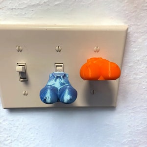May include: A light switch plate with three-dimensional, novelty light switch covers. One is blue and shaped like a pair of testicles, and the other is orange and shaped like a pumpkin. The light switch plate is white.