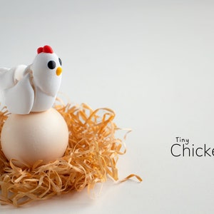 May include: A white clay chicken figurine perched on top of a white egg, nestled in a bed of straw. The text "Tiny Chicken" is written below the egg.