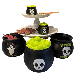 May include: Three black cauldron-shaped Halloween treat holders. One says "Here for the boos" with a ghost, one says "Witch Please" with a skull, and one has a skull and is filled with green candy.