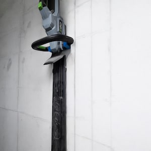 May include: A gray and black electric hedge trimmer with a green handle is mounted on a white wall using a blue and black wall mount.