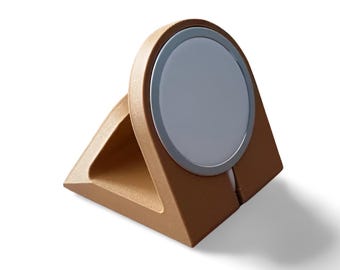 May include: A tan, triangular-shaped charging stand featuring a circular, light gray magnetic charging pad centered on the front face. The stand has a smooth, matte finish and a wide base, designed to hold a device at an angled position.