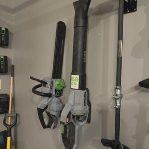 May include: A collection of outdoor power tools, including a leaf blower and a string trimmer, mounted on a wall. The leaf blower is gray and black with a green accent and a "650 CFM" label. The string trimmer is black and gray.