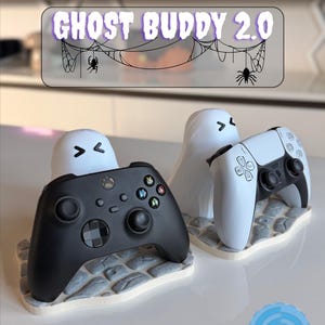 May include: Two ghost-shaped controller stands, one holding a black Xbox controller and the other a white PlayStation controller. The stands are on a gray stone-like base. The text "GHOST BUDDY 2.0" is above the stands.