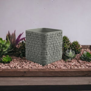 May include: A square gray planter with a stone-like texture. The planter is sitting on a wooden tray filled with small rocks and succulents.