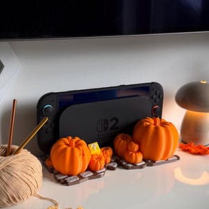 May include: A black Nintendo Switch console dock featuring a prominent number 2 logo, displayed on a brown brick-patterned base decorated with several orange pumpkins of varying sizes and a small yellow question mark block icon.