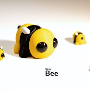 May include: A collection of yellow and black baby bee figurines with large black eyes and white accents. The word "Baby Bee" is written in black text below the figurines. The figurines are scattered on a white surface.