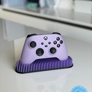 May include: A lavender purple Xbox controller with black buttons and a black Xbox logo is sitting on a lavender purple stand with black trim. The stand has a ribbed design.