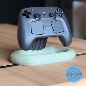 May include: A dark gray Steam controller resting on a light green, oval-shaped, ribbed display stand. The controller features a directional pad, two analog sticks, four action buttons labeled A, B, X, and Y, and two touchpads. The stand has a textured surface.