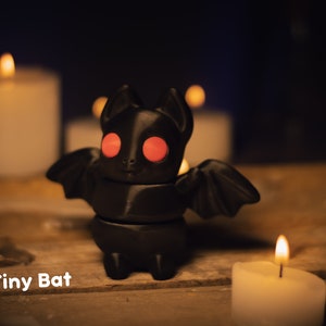 May include: A black 3D printed bat figurine with red eyes. The bat is standing on a wooden surface with white candles in the background. The text "Tiny Bat" is written below the bat.