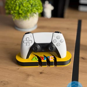 May include: A white and black wireless game controller rests on a yellow and black oval-shaped display stand. The stand features cutouts of a green triangle, blue circle, and pink square symbol. The text LUSPRINTS is visible in the bottom right corner.