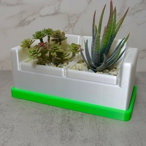 May include: A white sofa-shaped planter with a bright green base tray. The planter features two compartments holding small artificial succulents and decorative light-colored pebbles. The structure has a smooth finish and a rectangular design.