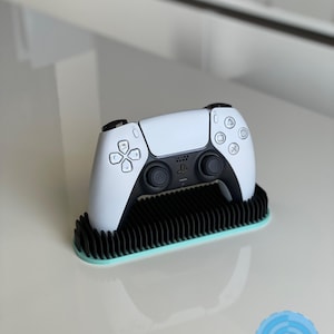 May include: A white PlayStation 5 controller is displayed on a black and teal stand. The stand has a textured surface.