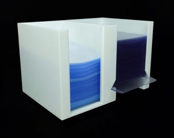 May include: A white plastic dual-compartment organizer holding two stacks of clear protective sleeves. The left stack features blue-tinted edges, while the right stack contains clear sleeves with a single sleeve extending outward from the base.