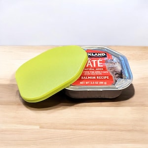 May include: A green plastic lid for a can of cat food. The can is silver and has a red label that says "KLAND ATÉ NATURAL JUICES FOOD FOR ADULT CATS GRAIN-FREE RECIPE SALMON RECIPE NET WT 3.5 OZ (99 g)".