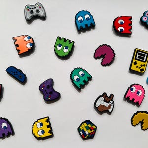 May include: A set of 16 colorful shoe charms featuring retro video game characters and icons. The charms include Pac-Man ghosts, a game controller, a game over sign, and a pixelated Rubik's Cube.