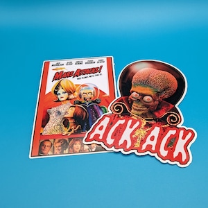 May include: A red and white sticker with the words "Ack Ack" in a bold, white font. The sticker features a close-up image of a Martian from the movie "Mars Attacks!" with a red and black costume and a large, green head.