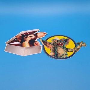May include: Two stickers featuring the character Gizmo from the movie Gremlins. One sticker shows Gizmo in a box, the other shows Gizmo with a menacing expression.