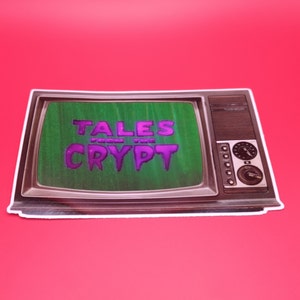 May include: A sticker featuring a vintage television with the words "TALES FROM THE CRYPT" in purple lettering on the screen. The TV has a brown frame and a green screen, set against a red background.