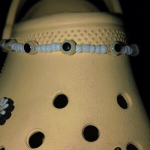 May include: A pair of yellow clogs with a black and white daisy charm and a black charm with the text "Bichora" in a barbed wire heart design. A white beaded bracelet with yellow and black eye charms is on the top of the clogs.