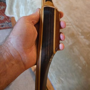 May include: A recurve bow riser constructed from laminated wood featuring alternating dark brown, black, and light tan vertical grain layers. The handle section includes a textured tan leather grip patch and visible white lettering printed on the side.