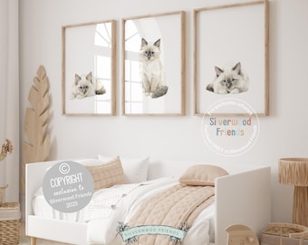 May include: Three framed cat art prints. Each print features a watercolor illustration of a Ragdoll kitten in different poses. The frames are light wood, and the artwork is displayed on a white wall. The room has a neutral color palette.