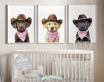 May include: Three framed prints featuring adorable puppies wearing cowboy hats and pink bandanas. The dogs are a chocolate lab, a golden lab, and a black lab. The artwork is displayed above a white crib.