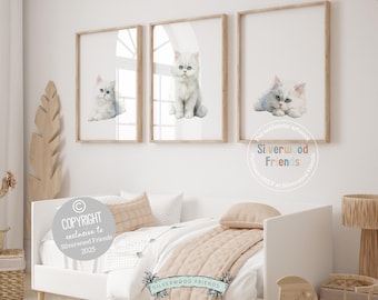 May include: Three framed prints featuring watercolor illustrations of white kittens with blue eyes. The kittens are in different poses. The frames are light brown, and the artwork is displayed on a white wall above a bed. The text "Silverwood Friends" is visible.