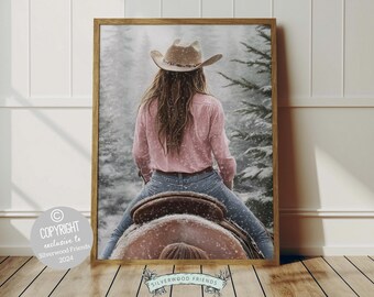 May include: A woman wearing a pink shirt and a brown cowboy hat rides a brown horse through a snowy forest.