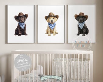 May include: Three framed prints featuring watercolor illustrations of puppies wearing cowboy hats and blue bandanas. The puppies are a chocolate lab, a yellow lab, and a black lab. The prints are displayed above a white crib.