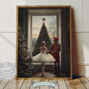 May include: A framed print depicting a ballerina in a white tutu standing beside a man in a red uniform. They are both gazing at a Christmas tree in the background. The scene is set in a room with a large window and a white wall.