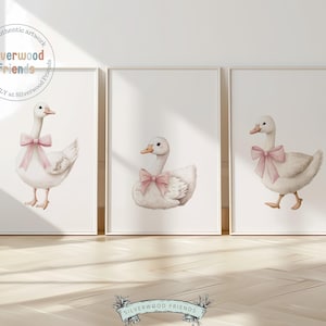 Op de afbeelding: Drie aquarel illustraties van eenden met roze strikken. Elke eend is afgebeeld in een andere pose. De illustraties zijn ingelijst in witte lijsten en worden getoond op een witte muur.